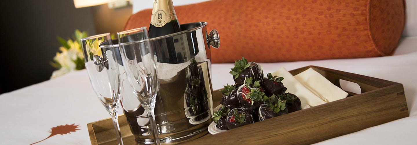 A bottle of champagne and two glasses sitting on a hotel guest bed with chocolate covered strawberries